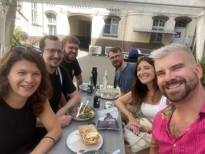 Whole team setting at a lunch table. Doing a selfie. Smiling into the camera. Some leftovers at the table. Factory Görlitzer Park coworking Space Innenhof lunch table
