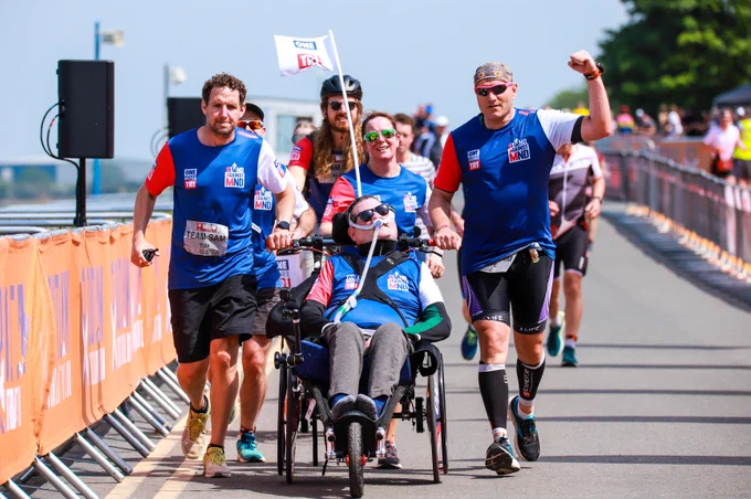 Sam Perkins completes the 26-mile running section of a triathlon in a specially-made wheelchair which holds his ventilator. He's being pushed by a group of friends called Team Sam. Image by: Ryan Sosna Bowd