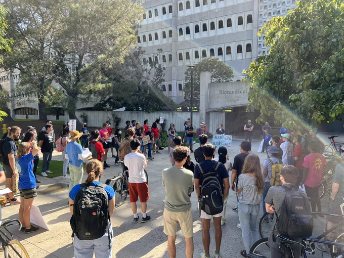 a crowd of protesters stand in a circle in a courtyard, holding signs and chanting