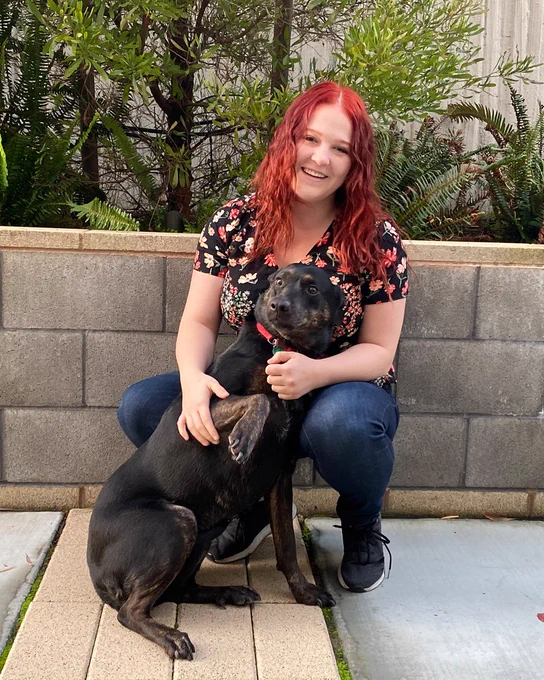 Nichole sits with a black dog in front of a cement wall with plants behind her.