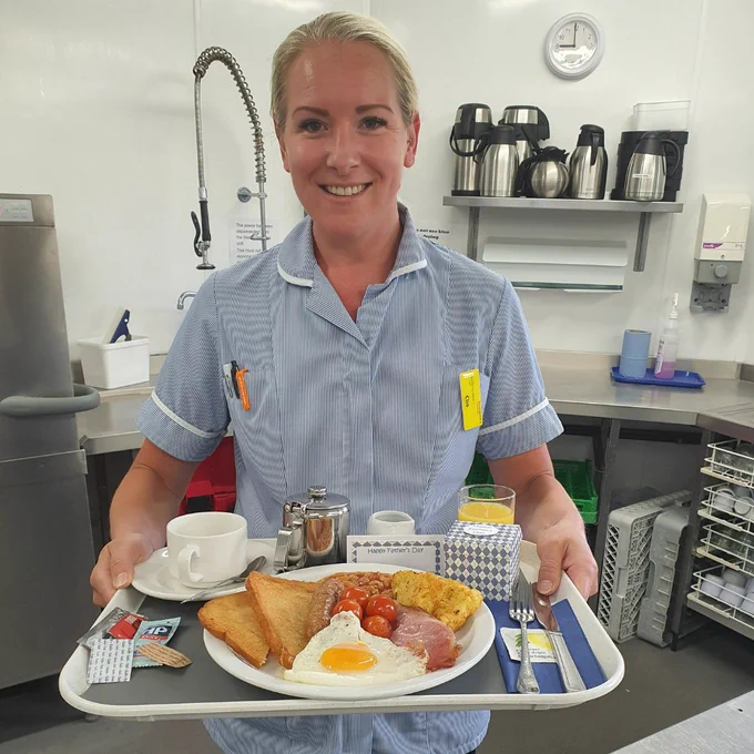 Nurse wearing blue and white striped uniform, smiling at the camera whilst holding a tray with full English breakfast, cup, teapot and orange juice.