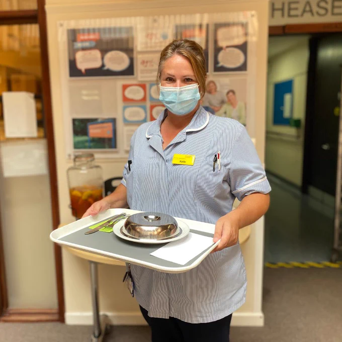 Nurse wearing blue and white striped uniform and face mask, looking at the camera whilst holding a tray with a covered plate and knife and fork.