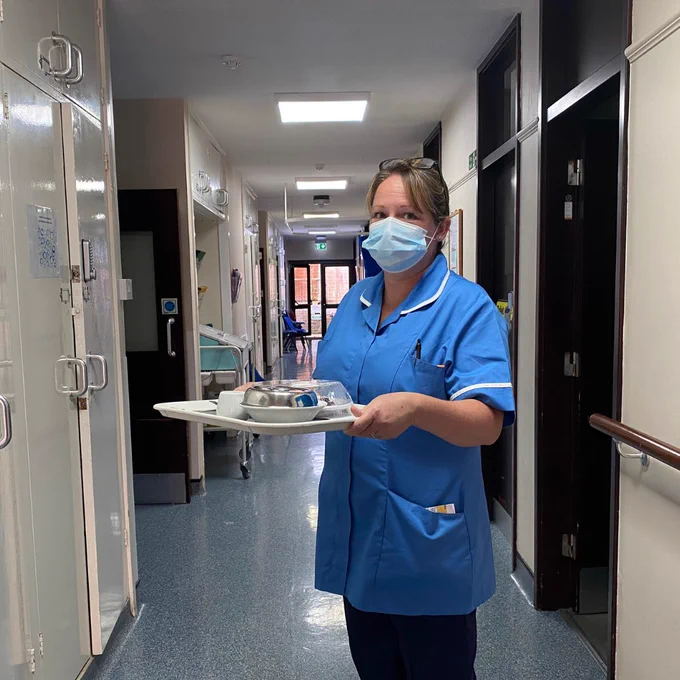 Nurse wearing royal blue uniform and face mask, looking at the camera whilst holding a tray with two covered plates and a small cup.