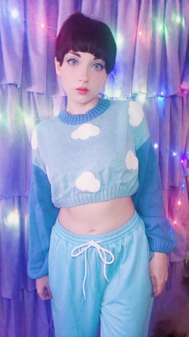 New looks straight at camera, posing in a blue outfit: a light blue crop-top sweater with dark sleeves and collar and little clouds adorning the torso, and light blue sweatpants. New has faint blue eyeshadow on with blue-grey circle lenses, soft blush, and a deep pink lip gloss. New's hair is short and black, with straight fairy-brows peeking out under the bangs. Behind them is a lavender curtain backdrop with multicolored fairy lights. 