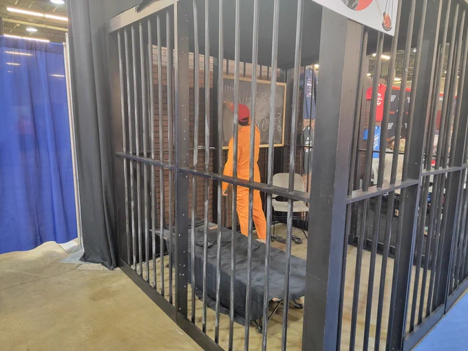 A man in an orange jumpsuit and MAGA hat writes something on a chalkboard in the middle of a very robust mock jail in the middle of a conference floor 