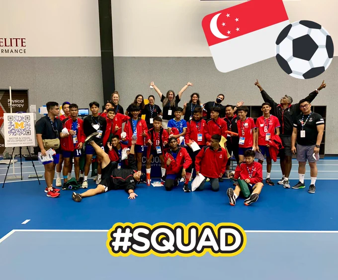 The University of Michigan Motor & Visual Development Lab team poses in black shirts with the Singapore men’s team in red shirts at the Special Olympics Unified Cup. Everyone is smiling or making a silly face, some have arms and legs extended, some are seated on the blue floor and some are standing against a gray and white background. Singaporean flag and soccer ball emojis are in the upper right corner, and #squad is written across the bottom in white with a black and yellow outline.