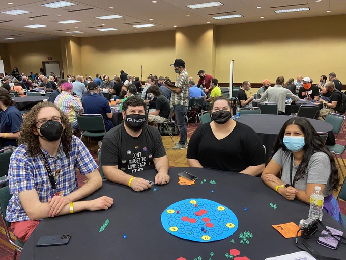 Foreground: colorful board game pieces on a round table. 4 playtesters sit around the table, smiling. The background is a busy bustle of playtest tables