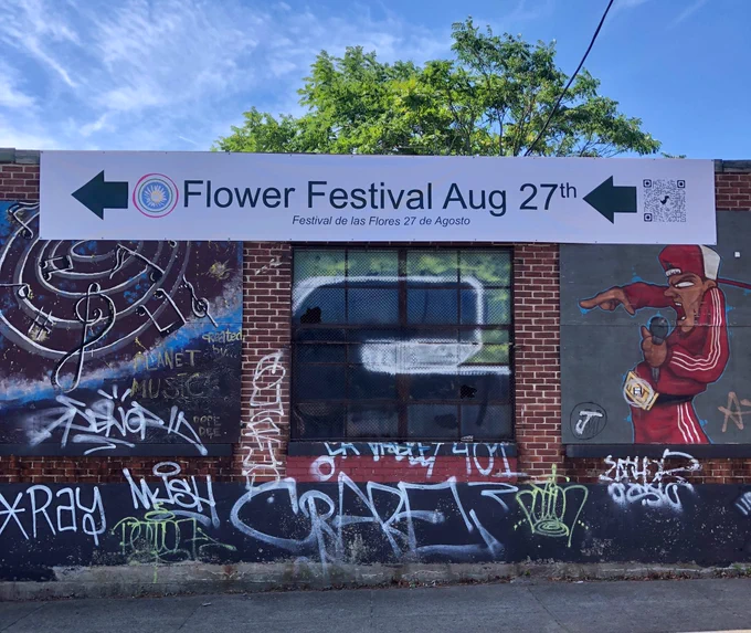 A one-story brick building that’s covered with graffiti has a sign at the top reading Flower Festival Aug 27