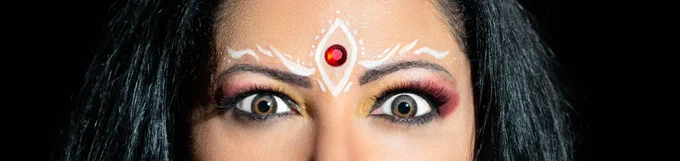 Photo by Devika Bilimoria  Image Description A woman’s eyes stare directly ahead. They are a piercing grey/brown with yellow and red eyeshadow. The image is a close-up on her eyes, with hair framing her face and you can only see blackness around her