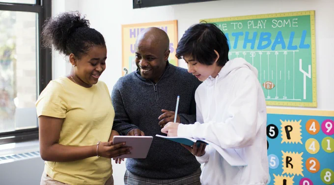 Photograph of a black bald teacher in a gray-blue sweater helping two teenage students: a black female student with her hair in a high bun wearing a yellow shirt and an asian male student with ear-length hair and a white hoodie. They are in a math, working on a problem with pencils and paper between them as they stand.