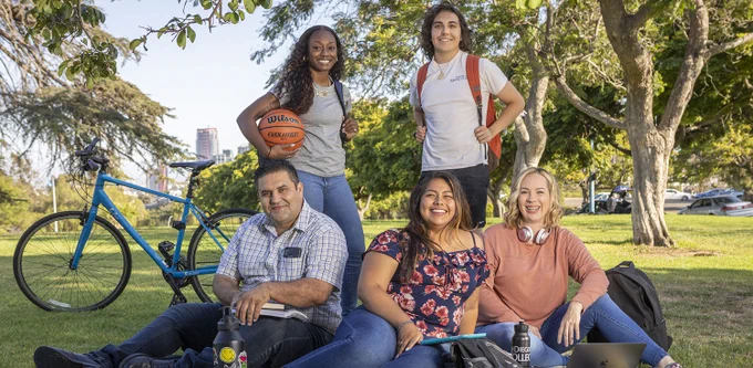 A male and two female students sit on the grass surrounded by books and a laptop. A female student stands behind them holding a basketball and a male student stands next to her wearing an orange backpack. There is a blue bicycle and trees in the background. 