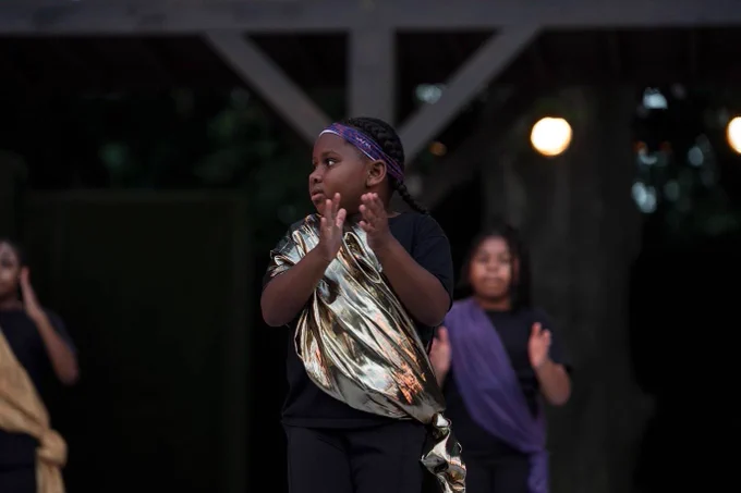 A young dancer performs as part of a step dance group. She wears a gold sash and is clapping.