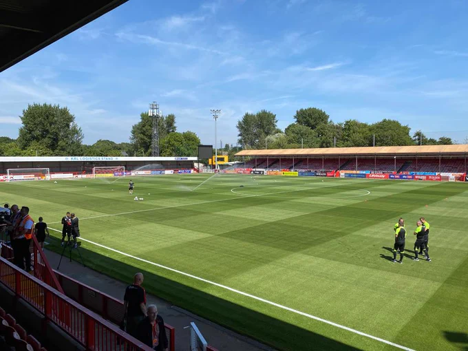 The photo shows the pitch at Broadfield stadium being prepared for today's match. Sprinklers are on and some players are warming up.