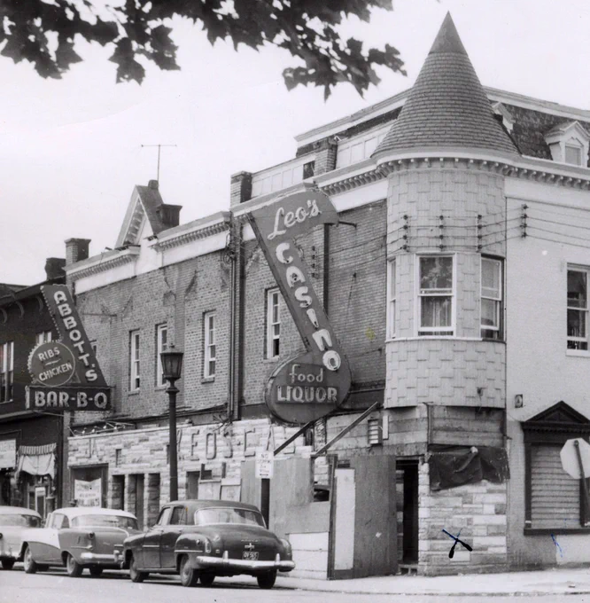 A corner commercial building faced with brick on the second story and formstone on the first story with a pointed turret on the near corner. Large signs are mounted on the second level: At center is a musical eighth-note shaped neon sign that reads “Leo’s CASINO food LIQUOR.” To the left is a checkmark shaped sign that reads “ABBOTT’S BAR-B-Q” with a circular seal-like medallion sign reading “Ribs—Chicken.” Three 1950s cars are parked in front and the leaves of a tree above the photographer appear at the top just inside the field of view.