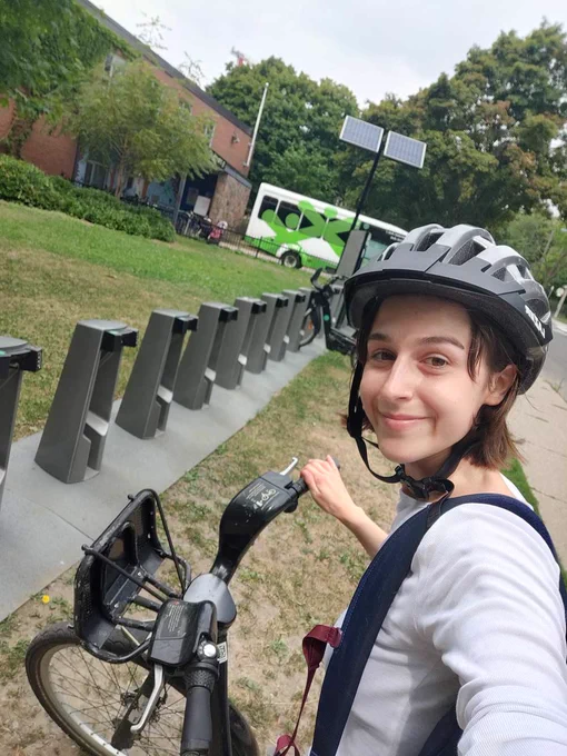 Bridget from TNGCS, wearing a bicycle helmet, smiles and gazes at the camera as she pulls a bicycle from a public rack. A shuttle bus sitting near some solar panels is visible in the background.