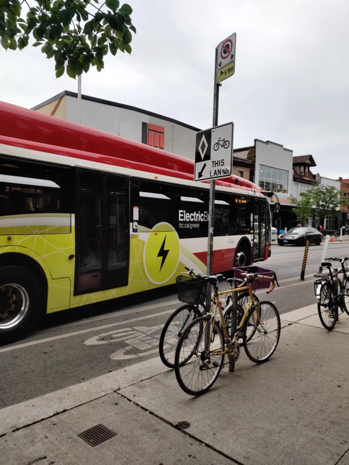 An electric transit bus drives past several bicycles leaning against poles and racks on a sidewalk.