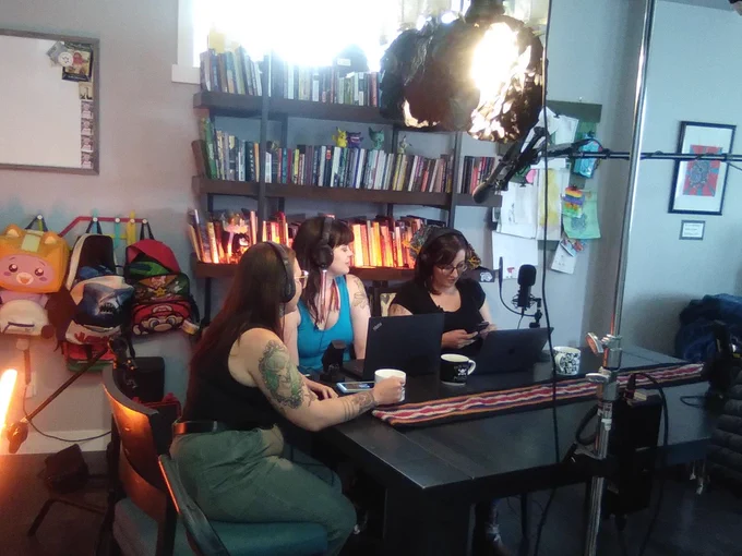 The three Book Women are sitting at a kitchen table; with computers and microphones.  A microphone is also above them being held by someone out of sight. 