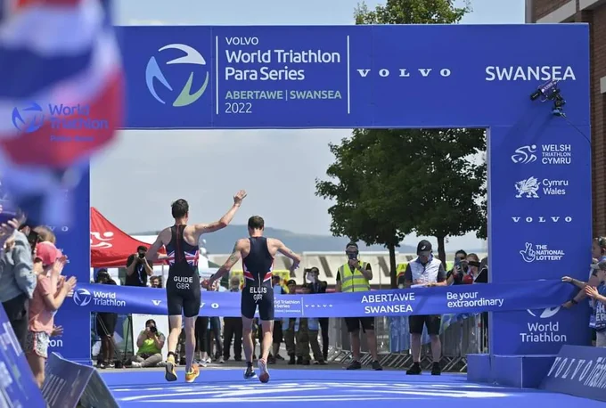 Dave and Luke crossing the finish line, Dave is about to run through the finish tape and Luke has an arm up being Dave celebrating. There is a big blue finish archway with world triathlon branding in and the crowd is out in their numbers! The photo is taken from behind Dave and Luke 
