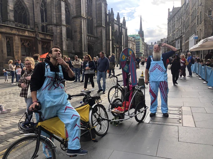 Two people wearing light blue dungerees with rhubarb pink geometric line pattern decals are with a black and yellow tandem bicycle with a light blue and rhubarb pink self built bicycle wheel of fortune they’ve called a selectatron attached to the back of it. Location is somewhere on the crowded royal mile in Edinburg during edinburgh festival fringe 2022. Person sat at the front of the tandem is pondering whilst another is stood saluting.
