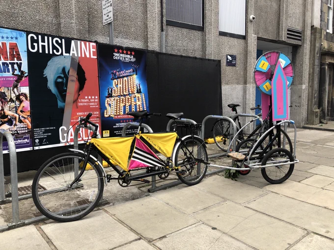 Black framed tandem decorated with triangles of yellow and pink material tied between gaps in the bike frame and a Bike trailer with a light blue and pink wheel of fortune attached. Edinburgh fringe bill boards in the background. Pavement with grey pavement slabs in foreground