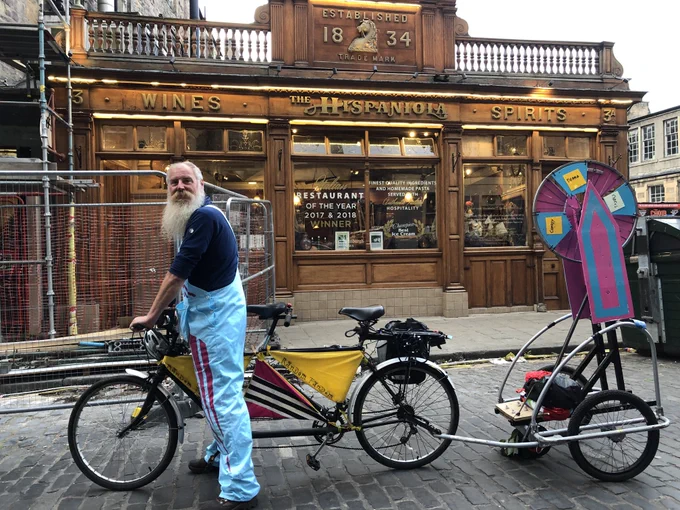 Man with light grey almost white beard wearing light blue dungerees is stood astride a black framed tandem bicycle decorated with triangles of material tied in between the gaps in the frame. A light blue and rhubarb pink selfbuilt bicycle wheel of fortune is on a bicycle trailer attached to the back. A wood fronted with large windows called the Hispaniola is in the background. Dark grey cobbles on the road surface.