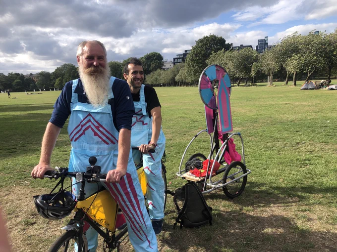 Two people on a tandem bicycle in a green treelined grassy park. one in foreground has a long light grey full beard the other on the backseat has short black hair no beard. Both wearing light blue dungarees with deep pink geometric line pattern decals. A bike trailer is attached has a tall light blue and pink wheel of fortune contraption created from a bike wheel and metal legs from a desk they are calling a selectatron with a large arrow pointing to a segment on the wheel