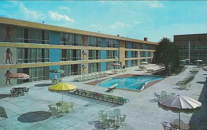 Courtyard view of Sahara Motor Hotel. An arrow-shaped swimming pool with low diving pool is in the center of a concrete courtyard and surrounded by rows of lounge chairs. Widely spaced multicolored umbrellas over tables and chairs line the perimeter of the courtyard. Egyptian figures are painted on the walls of the motel at regular intervals on all three floors. There is a single tree beyond the pool.