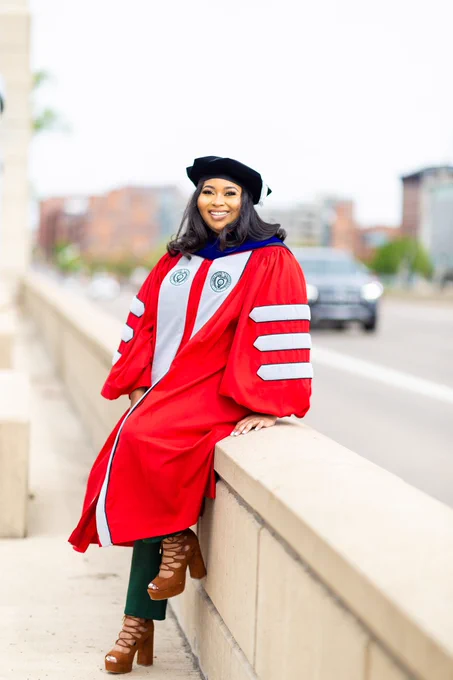 A Black woman sitting on a ledge on a walkway. She is wearing red and grey doctoral regalia.