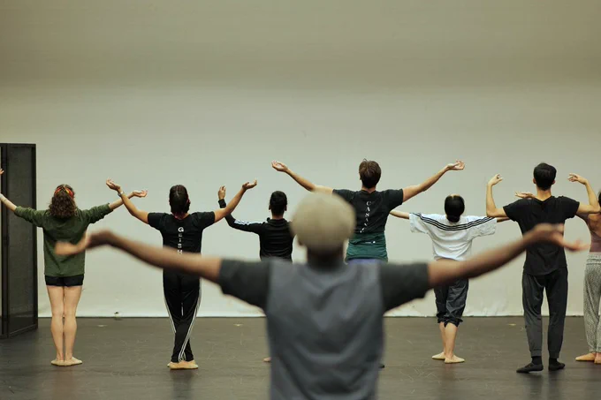 Northern Ballet dancers are in rehearsal, stood in a line with their backs to the camera. Their arms are out to the side with their hands turned upwards towards the ceiling. Photo by Mthuthuzeli November.