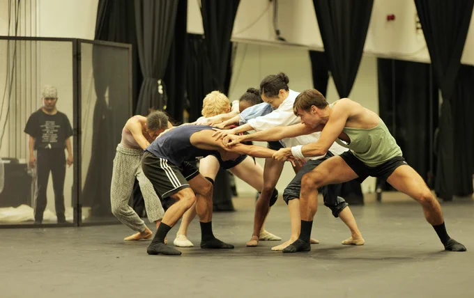 Northern Ballet dancers are in rehearsal. One dancer is crouched, looking down to the floor. They are surrounded by five dancers who have their hands on his shoulders and arms. Photo by Mthuthuzeli November.