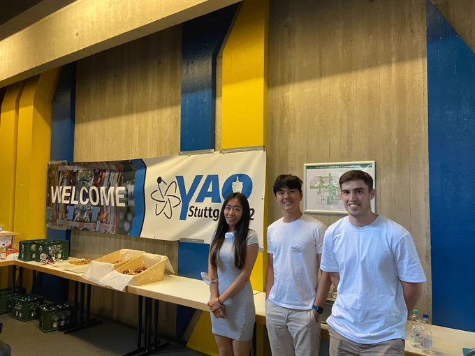 Three people stand smiling in front of a long table with some baskets of snacks and a banner reading "Welcome YAO Stuttgart '22"