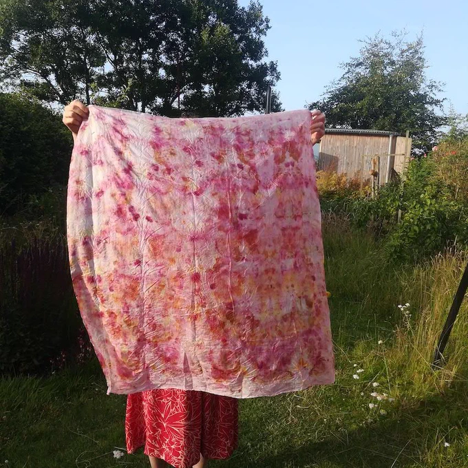 Person In a red floral dress holds up a sheet by the corners. The sheet is approximately 1.5m x 1.5m and has been dyed using the bundle dye technique and has beautiful patterns on it in pink and orange. The person stands in a garden with wild flowers. a shed stands at the end of the garden.