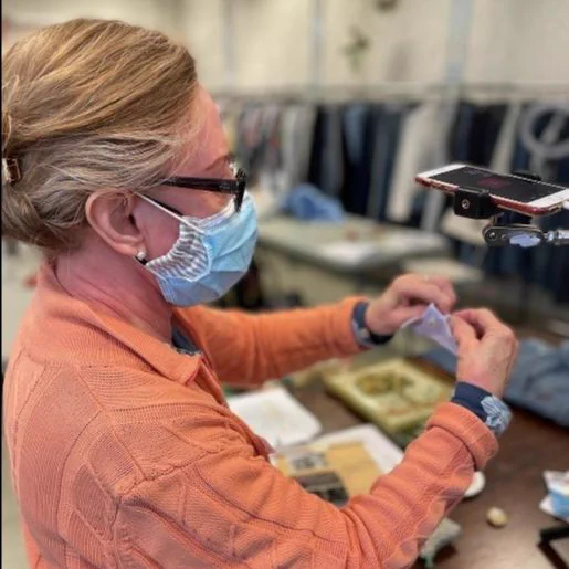 Mending demonstrator Jannette, wearing a mask and glasses, with her blonde/gray hair pulled back, shows a mending technique as it is being recorded on a phone. Racks of clothing and a table are in the background. Jannette will lead a mending workshop at TNG Community Services' Central Neighbourhood House location at 349 Ontario Street, Toronto on August 11th from 11 am to 1 pm.