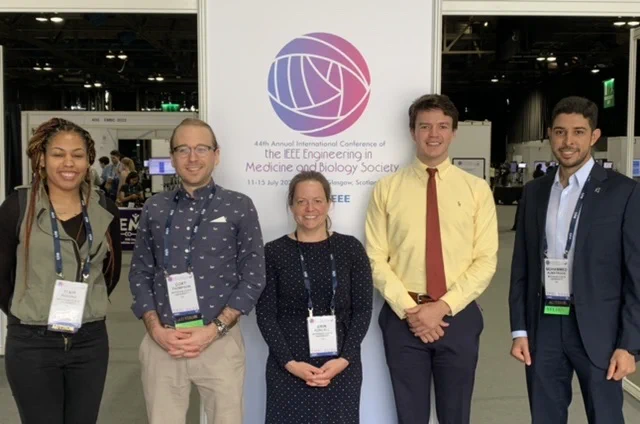 Photo of five people standing side by side in front of a poster for the IEEE conference