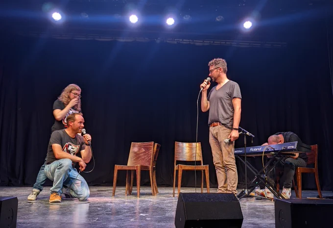 Photo of four men performing onstage - three with microphones in various poses, one at a keyboard.