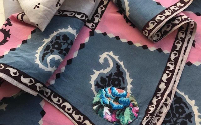 A block printed piece of fabric with paisley pattern in black around the borders with squares of blue and pink with paisley patterns block printed has been mended with a brightly coloured floral patch. It is a delicate fabric made of silk that has been hand stitched to repair a rip or hole in the fabric