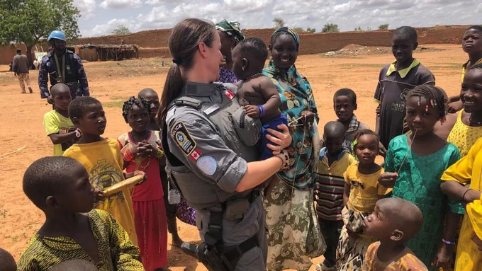 A Canadian woman police officer stands holding a Malian baby, while a dozen children stand around her, with parents on the outskirts of the group smiling, outside in Mali.