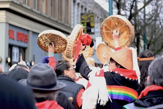 A photo of a group of Indigenous women on a city street walking away from the camera holding hand drums. They are wearing black and red, and one is wearing a cedar-woven hat and a rainbow ribbon shirt.