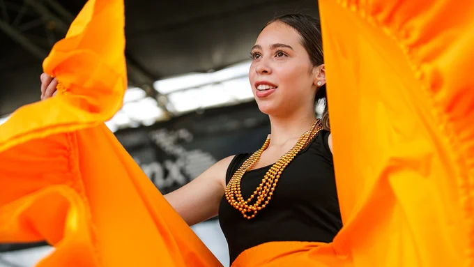 A young woman holds up the sides of her vibrant orange skirt while performing a dance. She wears orange beaded necklaces over a black shirt.