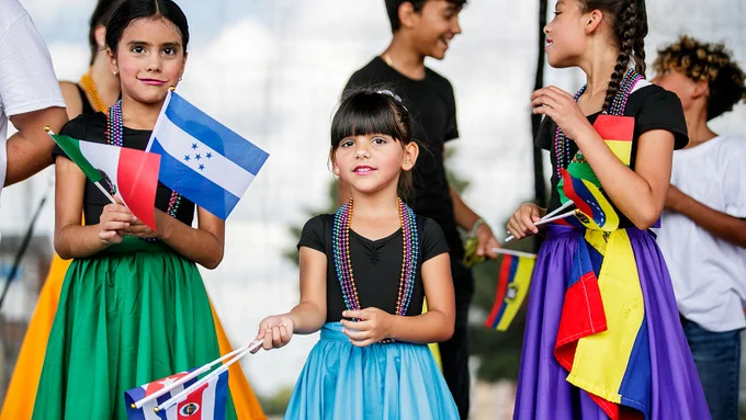 A group of children wearing beaded necklaces and brightly colored skirts wave flags from Latin American countries.