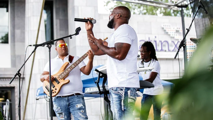 Three members of a band wearing white shirts and jeans play guitar, sing and play a keyboard while performing on an outdoor stage.