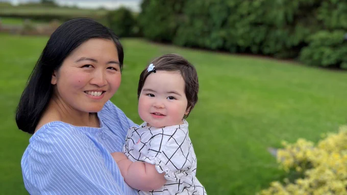 Laura holding baby Chloe in her arms. They're both smiling, looking at the camera and standing in a garden.