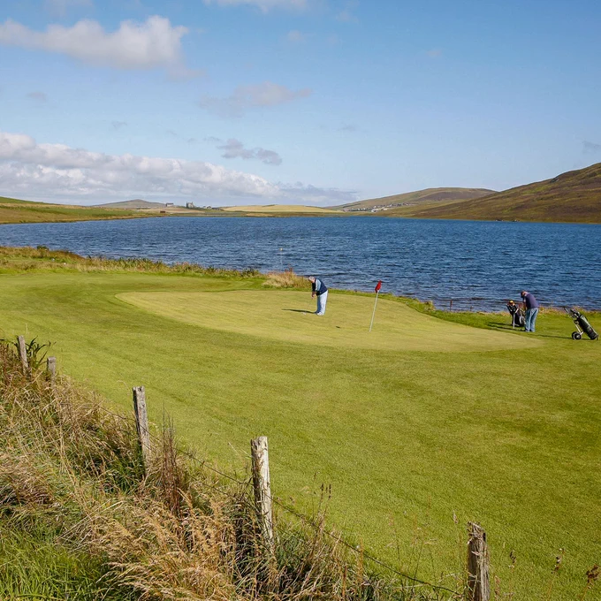 Image shows two golfers putting on the green on the 9-hole course at Asta Golf Club, beside Loch of Asta, near Scalloway, Shetland
