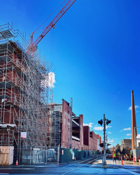 Construction site of the Met Warehouse in Cambridge, MA, seen from the street. Clear blue skies, scaffolding, workers in the distance. 