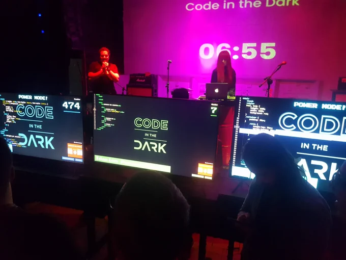 Fotografia em ambiente escuro. Ao fundo um telão em rosa com um texto centralizado 
"Code in the Dark

06:55"

A frente do telão, cuas pessoas, uma  com um microfone. No primeiro plano, 3 televisões apresentando códigos em HTML"