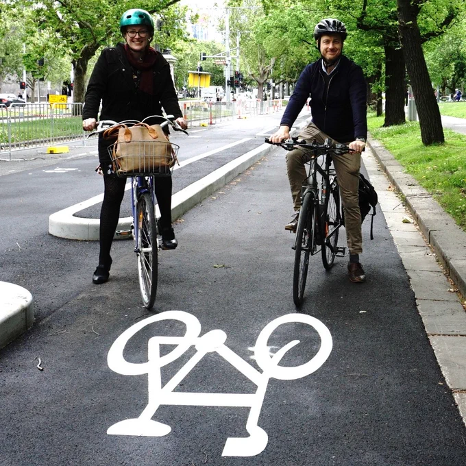 Katherine and Sam Hibbins sit on bikes in a newly-completed separated bike lane. There is a bike symbol painted on the bitumen in front of them, and the lane is separated from the main roadway by a wide curb. There are green grass and trees in the background on surrounding medians