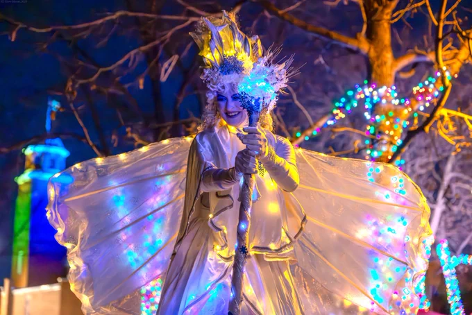 Stilt Walker dressed as Winter fairy standing in front of tree lit up with fairy lights