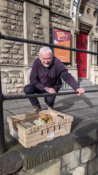 A staff member smiling whilst kneeling down next to a crate of donated pumpkins on the floor. He is resting his hand on the railings and it's a sunny day.
