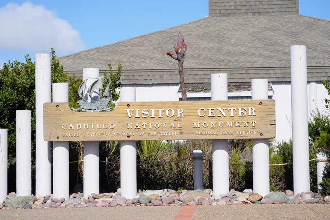 A sign in the center of the photo reads “Visitor Center, Cabrillo National Monument, United States Department of the Interior, National Park Service.” A large greenish-red stalk is visible behind the sign.