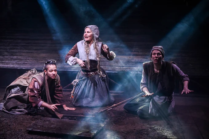 Three actors dressed in long skirts with rope belts kneeling on a dark set highlighted with pinpoints of light. They floor looks like soil and their clothes are grubby. 
